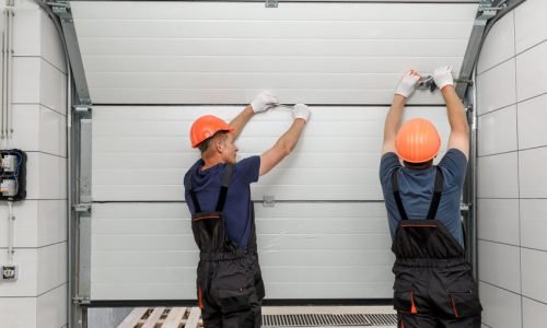 Entretien d’une porte sectionnelle dépannage rideaux métalliques Deux Techniciens en casque orange qui entretiennent une porte de garage sectionnelle en PVC Blanc dépannage rideaux métalliques